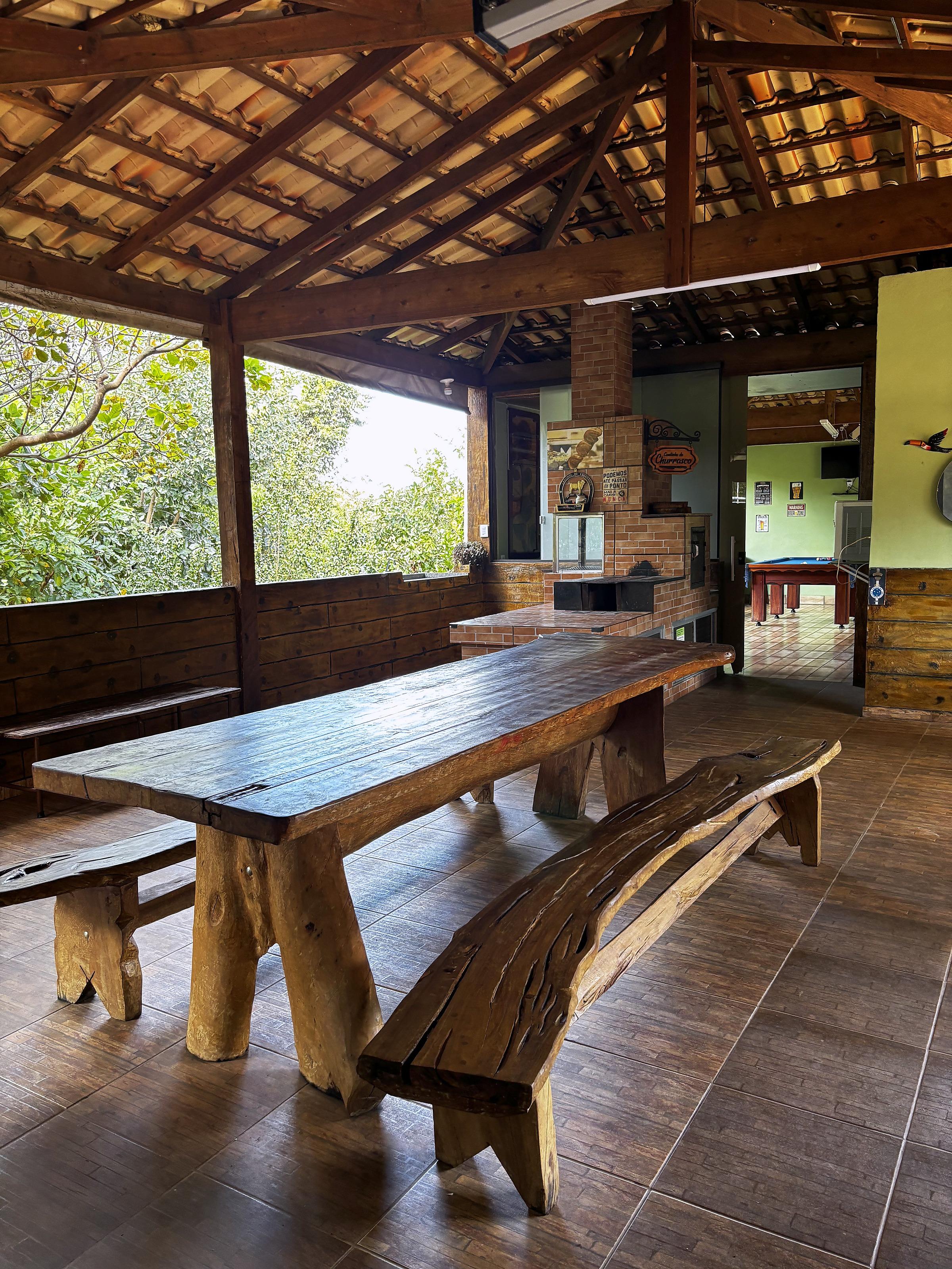 Rancho coberto com mesa rústica de madeira e churrasqueira ao fundo, vista para o jardim